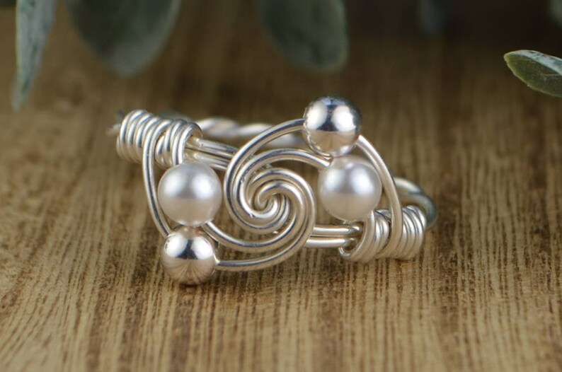 May include: A silver-toned wire ring with a spiral design, featuring three pearl-like beads and two silver beads. The ring is set against a blurred wooden background, highlighting the intricate wirework and the contrast of the shiny beads.