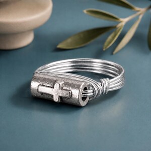 May include: A silver-toned ring constructed from multiple thin wire bands that wrap around a central cylindrical bead. The bead features a raised cross emblem on its front surface, and the wire bands are coiled tightly at the sides of the bead.