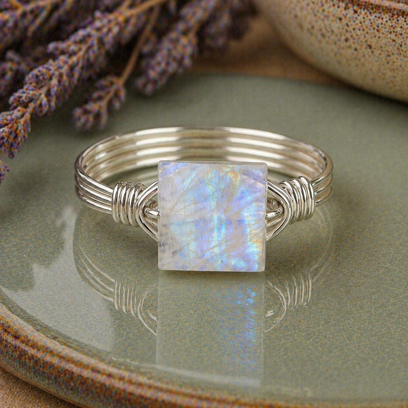 May include: A wire-wrapped ring featuring a square-cut moonstone with iridescent blue flashes. The band is constructed from multiple strands of silver-colored wire that coil around the base of the square gemstone, securing it in a central position.