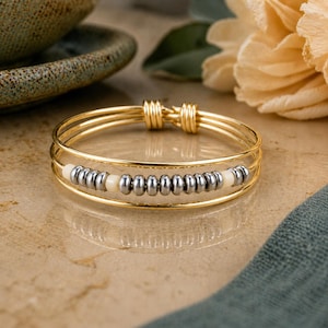 May include: A gold-toned wire bangle bracelet featuring three parallel bands. The centre band is adorned with a row of small, metallic silver-coloured beads interspersed with two smooth, cream-coloured cylindrical beads at each end of the row.