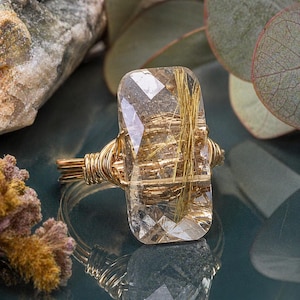 May include: A ring featuring a rectangular, faceted rutilated quartz gemstone with visible golden needle-like inclusions. The stone is secured by a gold-toned wire-wrapped band that coils around the base of the gemstone to hold it in place.