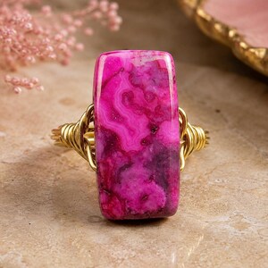 May include: A rectangular ring featuring a vibrant pink agate stone with swirling patterns. The stone is set in a gold-colored wire wrap design. The ring is a statement piece, showcasing a unique gemstone.