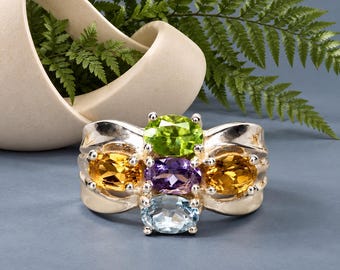 May include: A silver ring featuring a cluster of four oval-cut gemstones. The stones include a bright green peridot at the top, a central purple amethyst, a light blue topaz at the bottom, and two golden yellow citrines on the left and right sides.