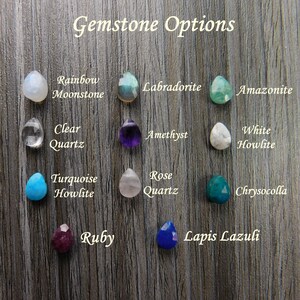 Pode incluir: Uma sele&ccedil;&atilde;o de pedras preciosas em forma de gota, incluindo Rainbow Moonstone, Labradorite, Ametista e Quartzo Rosa, &eacute; apresentada sobre um fundo de gr&atilde;o de madeira. O texto "Gemstone Options" est&aacute; no topo. Outras pedras incluem Clear Quartz, Turquesa Howlite, Rubi e Lapis Lazuli.