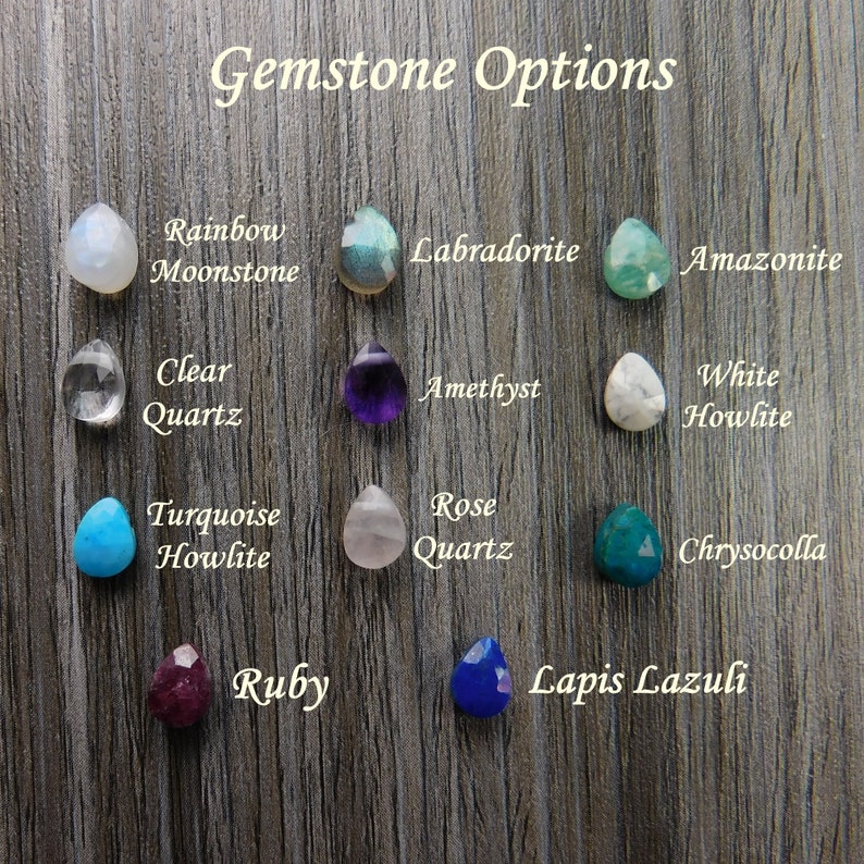 Pode incluir: Uma sele&ccedil;&atilde;o de pedras preciosas em forma de gota, incluindo Rainbow Moonstone, Labradorite, Ametista e Quartzo Rosa, &eacute; apresentada sobre um fundo de gr&atilde;o de madeira. O texto "Gemstone Options" est&aacute; no topo. Outras pedras incluem Clear Quartz, Turquesa Howlite, Rubi e Lapis Lazuli.