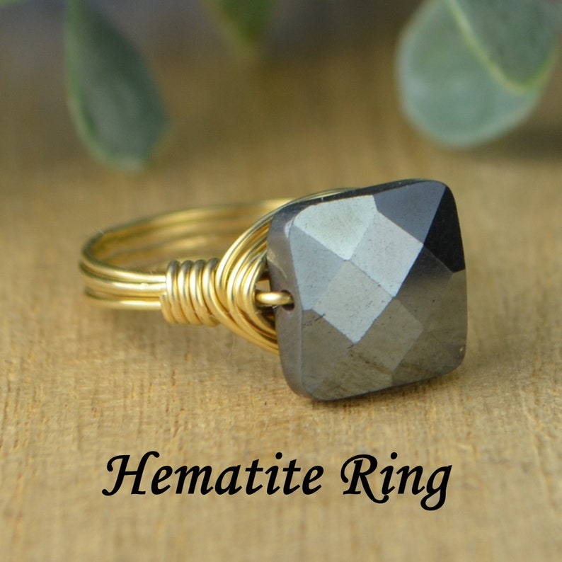 May include: A gold-toned wire-wrapped ring with a square, faceted hematite stone. The ring features the text "Hematite Ring" in a cursive font, set against a wooden background. The hematite stone is a dark gray color.