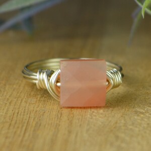 May include: A silver wire-wrapped ring featuring a square, peach-colored gemstone. The gemstone is faceted, catching the light. The ring is set against a wooden surface, showcasing the jewelry's design.