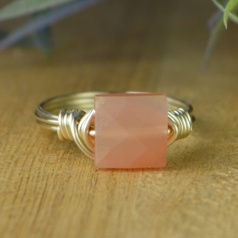 May include: A silver wire-wrapped ring featuring a square, peach-colored gemstone. The gemstone is faceted, catching the light. The ring is set against a wooden surface, showcasing the jewelry's design.
