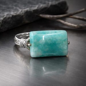 May include: A silver wire-wrapped ring featuring a rectangular, light teal gemstone. The stone has a marbled appearance with white and darker teal inclusions. The ring is set against a metallic surface with a blurred background.