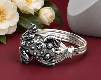 May include: A silver-toned metal bangle bracelet featuring a textured frog ornament on the front. The bracelet is constructed from multiple thin, coiled wire bands that wrap around to form a circular shape with a polished, metallic finish.