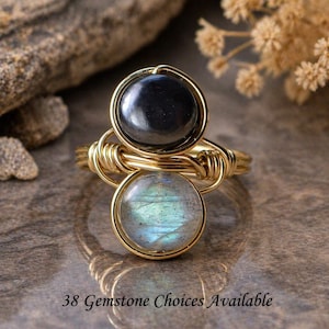 May include: A gold-toned wire-wrapped ring featuring two round gemstones. The top stone is black, and the bottom stone is a light blue with iridescent qualities. The ring has the text "38 Gemstone Choices Available".