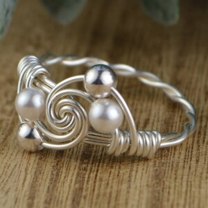 May include: A silver-toned wire ring with a spiral design and small, round, white pearls. The ring features a twisted band and is set against a wooden surface.