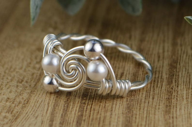 May include: A silver-toned wire ring with a spiral design and small, round, white pearls. The ring features a twisted band and is set against a wooden surface.