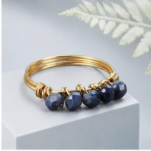 May include: A gold-toned wire ring featuring five faceted, deep blue beads. The beads are individually wrapped with thin gold wire along the lower section of the band, creating a textured, layered appearance with a polished metallic finish.
