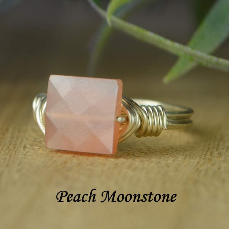 May include: A silver wire-wrapped ring featuring a square, peach-colored moonstone. The gemstone has a faceted surface, catching the light. The ring is set against a blurred wooden background with the text "Peach Moonstone".
