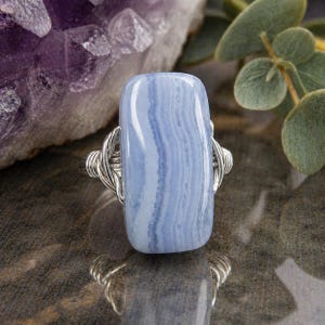 May include: A rectangular blue lace agate gemstone ring with a silver wire wrap. The stone has light blue and white bands. The ring is set against a blurred background of a purple crystal and green leaves.