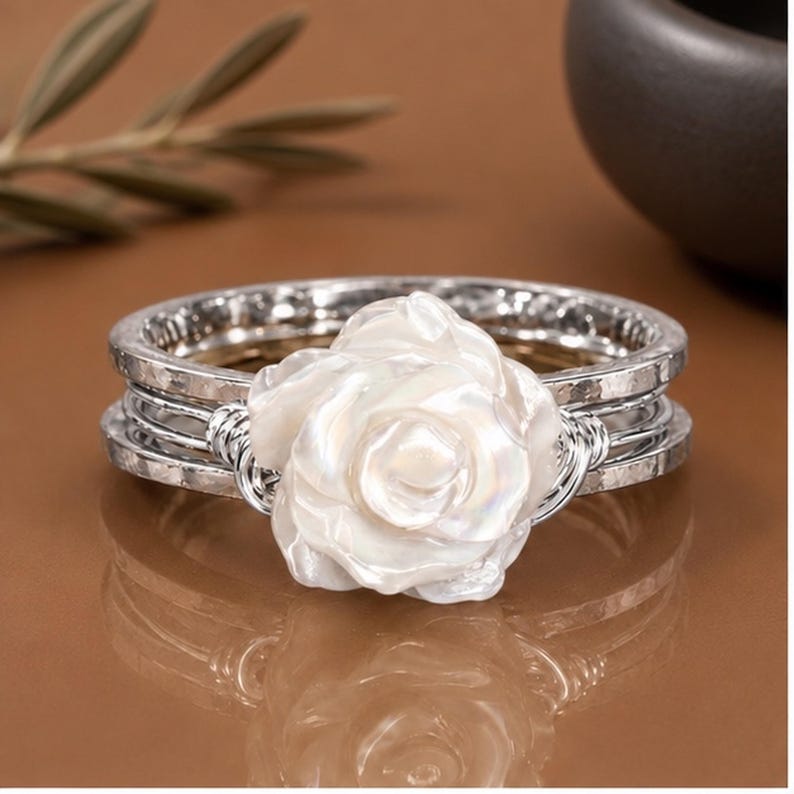 May include: A silver-toned ring featuring a central white, iridescent, carved flower element. The band consists of multiple thin, hammered silver-toned wires that are wrapped together at the sides to secure the floral centrepiece in place.