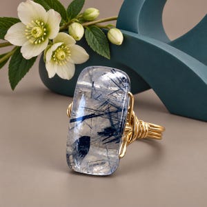 May include: A ring featuring a large, rectangular, clear rutilated quartz gemstone with dark blue needle-like inclusions. The stone is secured by a gold-toned wire-wrapped band that coils around the sides of the gemstone setting.