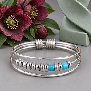 May include: A silver bangle bracelet featuring three parallel bands connected by two silver coils. The center band is adorned with a row of smooth silver beads interspersed with two bright turquoise blue cylindrical beads.