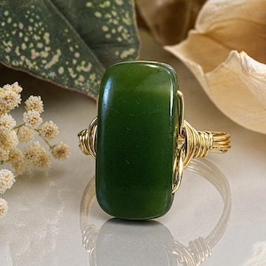 May include: A handcrafted ring featuring a rectangular, deep green jade stone. The stone is encased in a gold-colored wire wrap design. The ring is displayed on a reflective surface, showcasing the intricate wirework and the jade's polished finish.