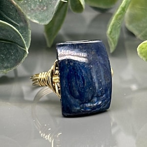 May include: A handcrafted ring featuring a rectangular, deep blue stone. The stone is set in a gold-colored wire wrap design. The ring is a unique piece of jewelry.