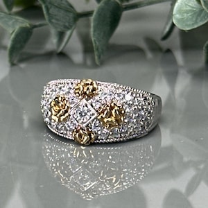 May include: A silver-toned ring with a central square diamond and surrounding smaller clear stones. Gold-toned floral accents are interspersed throughout the design, creating a luxurious and detailed appearance. The ring is set against a reflective surface.