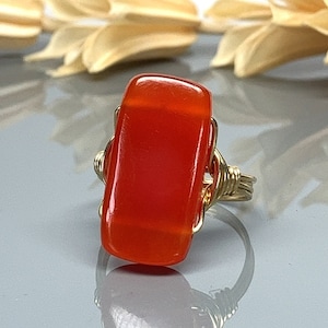 May include: A handmade ring featuring a rectangular, orange-red gemstone set in a gold-colored wire wrap. The gemstone has a polished, glossy finish. The ring's band is also gold-colored.