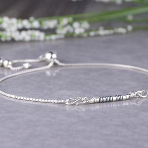 May include: A delicate silver bracelet with a sliding clasp. The bracelet features a fine chain and a decorative section with small silver and dark grey beads. The design is simple and elegant, suitable for everyday wear.