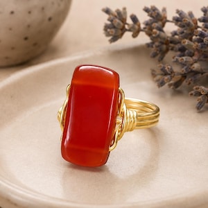 May include: A ring featuring a rectangular, smooth, deep orange-red carnelian gemstone set in a gold-colored wire-wrapped band. The wire is coiled around the sides of the stone, creating a secure setting for the polished, vibrant red-orange cabochon.
