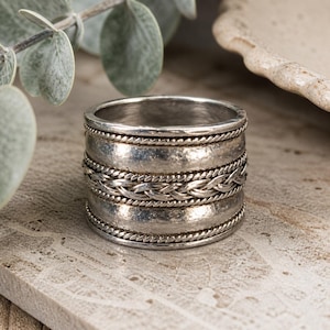 May include: A chunky silver ring with a textured finish and braided detailing. The ring has multiple bands with a hammered metal effect, framed by fine rope-like borders. The design is striking and suitable for everyday wear. The ring's width is approximately 1.5 cm.