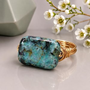 May include: A ring featuring a rectangular, polished turquoise stone with dark matrix veining. The stone is secured by a multi-strand gold-toned wire band that wraps around the sides of the gemstone, creating a layered, coiled effect on the shank.