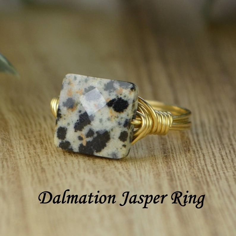 May include: A gold-toned wire-wrapped ring featuring a square dalmatian jasper stone. The stone has a speckled pattern of black, white, and tan. The ring is set against a wooden surface. Text on the image reads "Dalmatian Jasper Ring."