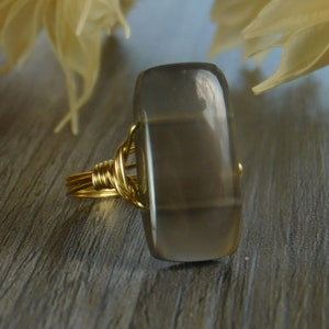 May include: A rectangular gray gemstone ring with a gold wire wrap. The gemstone is a light gray color with darker gray bands. The gold wire is wrapped around the gemstone and forms the ring band.
