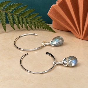 Pode incluir: Um par de brincos de argola em prata com pedras de labradorite facetadas em forma de gota. Cada pedra está presa ao fio de prata fino e circular por um enrolamento de fio de prata, exibindo os tons iridescentes de azul e cinzento da pedra.