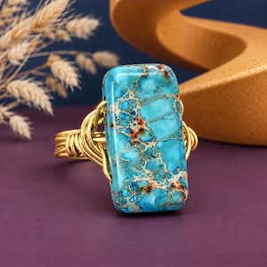 May include: A ring featuring a rectangular turquoise-colored stone with brown veining, set in a gold-toned wire-wrapped band. The polished stone has a smooth surface and is secured by multiple loops of gold-colored wire on each side of the setting.