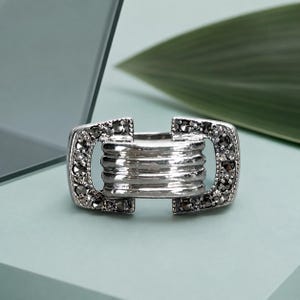 May include: A silver-toned ring featuring a central horizontal bar design with four parallel ridges. The two rectangular ends of the ring are embellished with small, dark, faceted stones set in a metallic frame, creating a structured appearance.