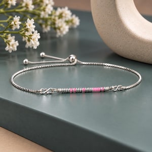 May include: A silver box chain bracelet featuring an adjustable sliding clasp and a central horizontal bar. The bar is decorated with a series of small, alternating silver and pink cylindrical beads arranged in a linear pattern along the chain.