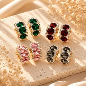 Puede incluir: Cuatro pares de pendientes de aro con tres piedras de corte redondo cada uno, rodeadas de pequeños cristales transparentes. Los juegos incluyen piedras verde oscuro, rojo oscuro, rosa suave y gris carbón, montadas en metal dorado.