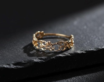 May include: A gold-colored ring with an intricate floral design. The ring features delicate leaves and swirling patterns, creating an elegant and ornate look. The ring is set against a dark, textured surface.