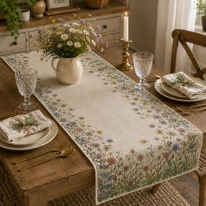 Può includere: Un runner da tavola rettangolare color beige con un bordo stampato di fiori di campo colorati, tra cui margherite, fiordalisi e fiori rosa. Il runner è posizionato su un tavolo in legno con una brocca in ceramica e bicchieri.