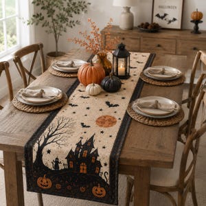 May include: A rectangular table runner featuring a printed Halloween scene with a haunted house, a large orange moon, bats, stars, and jack-o'-lanterns. The runner is made of a beige fabric with black borders and is displayed on a wooden dining table.