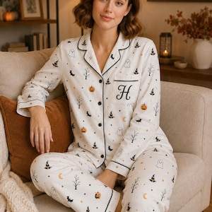 Könnte beinhalten: Ein weißer, langärmeliger Pyjama mit durchgeknöpftem Oberteil und passender Hose, verziert mit einem wiederkehrenden Halloween-Muster aus orangefarbenen Kürbissen, schwarzen Hexenhüten, weißen Geistern, Fledermäusen und kahlen Bäumen.