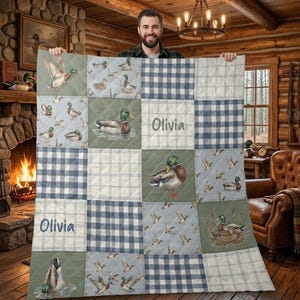 May include: A quilted blanket featuring a patchwork design with duck illustrations and blue plaid squares. The blanket has the name "Olivia" embroidered on it. The color palette includes shades of blue, green, and white.