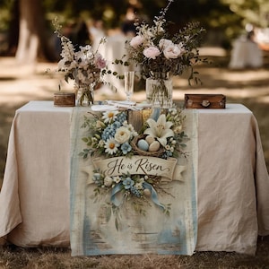 May include: A beige tablecloth with a floral table runner featuring the text "He is Risen." The runner has a nest with eggs, white lilies, and blue and white flowers. Vases of flowers and small wooden boxes are on the table.