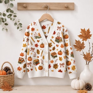 May include: A white V-neck cardigan featuring a repeating pattern of orange pumpkins, colorful autumn leaves, stacks of books, pencils, and small bows. The long-sleeved knit garment has a button-front closure with ribbed cuffs and a ribbed hem.