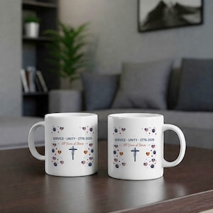 May include: Two white ceramic mugs with a handle, each featuring a design with the text "SERVICE UNITY 1776-2026 - 250 Years of Stories". The design includes a cross, hearts, and dots in red, white, and blue.