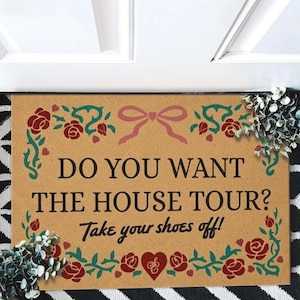 May include: A rectangular doormat with the text "DO YOU WANT THE HOUSE TOUR? Take your shoes off!" in black. The mat has a tan background with a pink bow and red roses and green vines. The mat is on a black and white patterned surface.