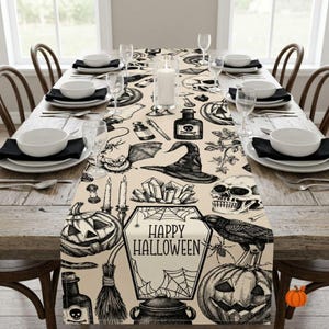 Op de afbeelding: Een beige tafelloper met een zwarte inktprint van Halloween-motieven, waaronder pompoenen, een schedel, een heksenhoed, een raaf, kristallen en een centraal doodskistvormig kader met de tekst Happy Halloween omringd door spinnenwebben.