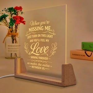 May include: A lighted acrylic sign with a wooden base displays the text "When you're MISSING ME, just turn on this light and you'll feel my Love SHINING THROUGH no matter the miles BETWEEN US." A vase of flowers is in the background.