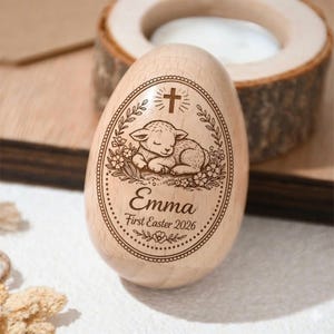 May include: A wooden egg-shaped ornament with a laser-etched design. The design features a sleeping lamb, a cross, floral elements, and the name "Emma" with "First Easter 2026". The egg is light brown and sits on a wooden surface.